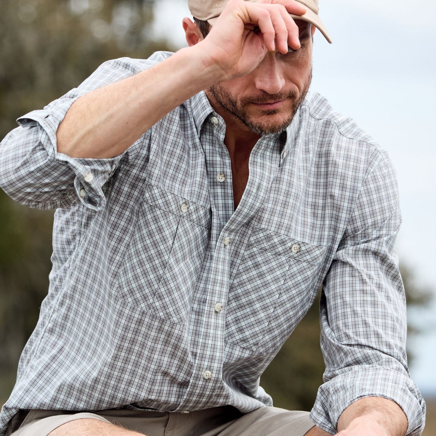 Man in a Tom Beckbe Tidewater Shirt adjusts his hat outdoors, partially covering his face.