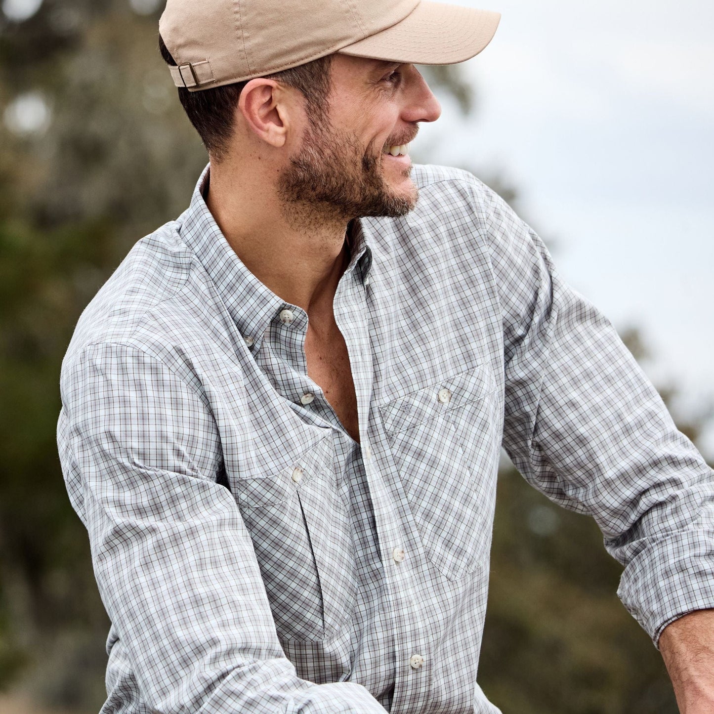 A man wearing a Tom Beckbe Tidewater Shirt and beige cap smiles, looking to the side outdoors.