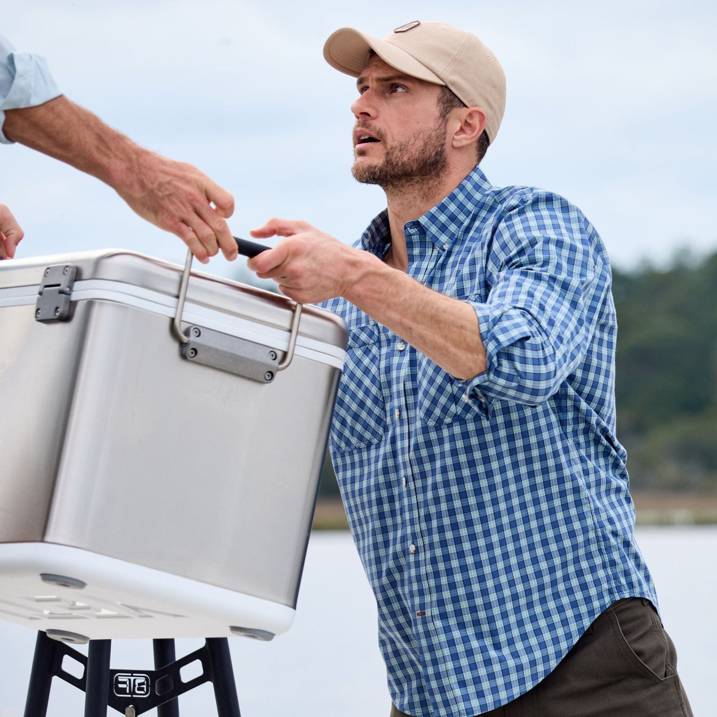 A man in a Tom Beckbe Tidewater Shirt hands a cooler to another person near water.