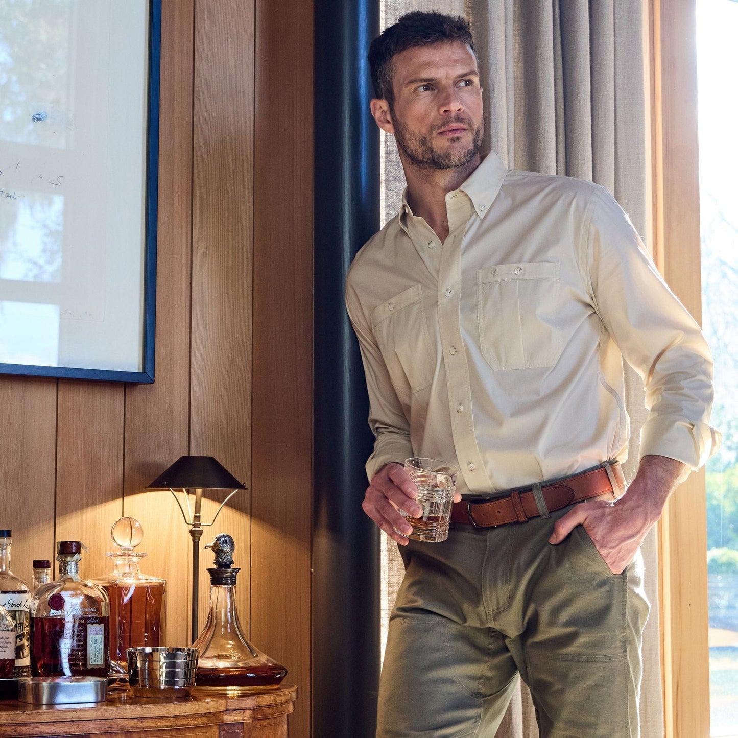 A man in a beige Tom Beckbe Tidewater Shirt stands by a window, holding a glass, with bottles and a lamp nearby.