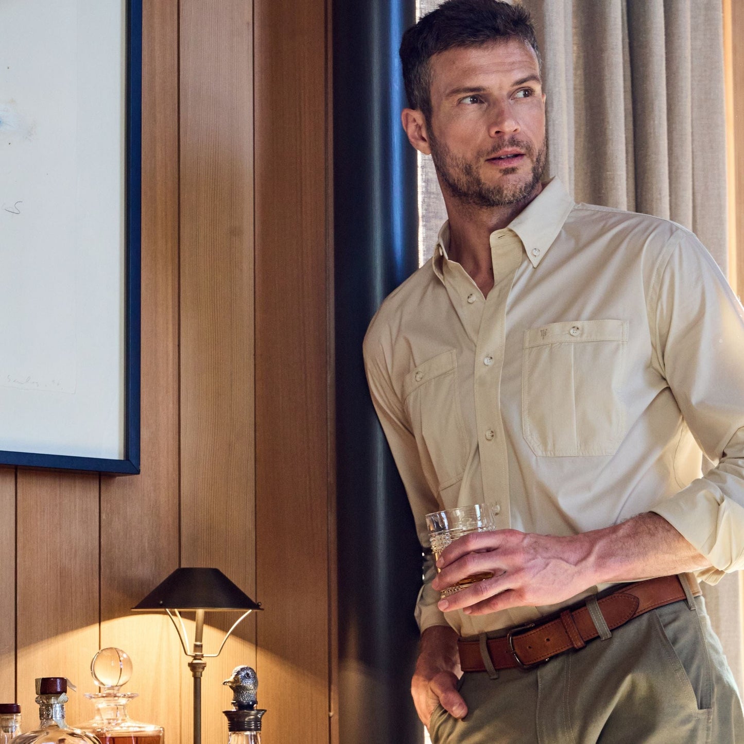 Man in a Tom Beckbe Tidewater Shirt holds a drink, standing by a wooden wall with framed art and a lamp behind him.