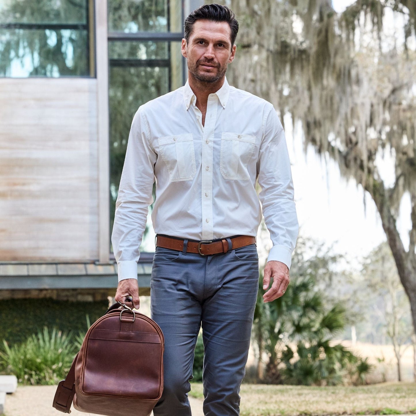 A man in a Tom Beckbe Tidewater Shirt and gray pants strolls outside with a brown bag under tree shade.