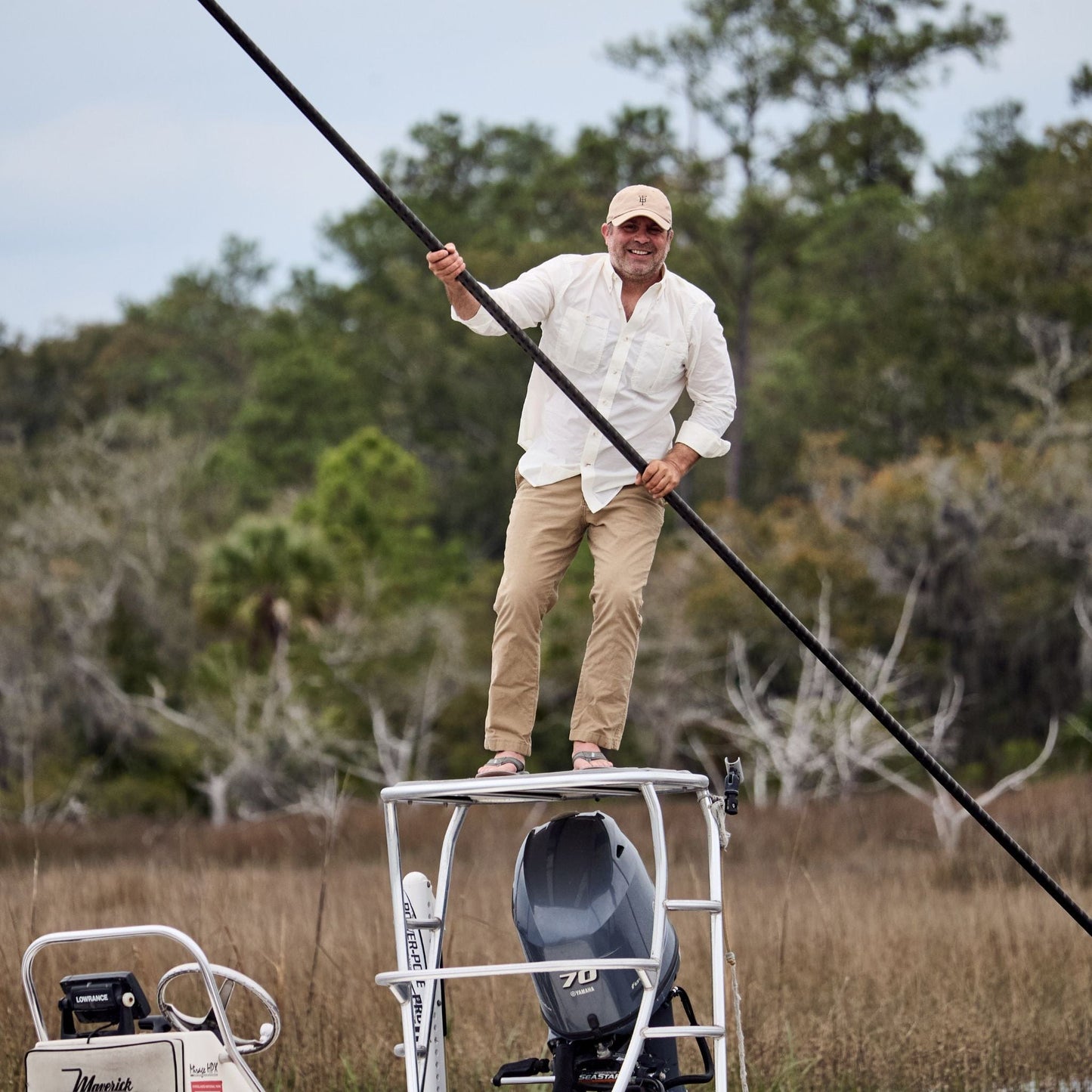 Man in a hat atop a boats tower, wearing a Tom Beckbe Tidewater Shirt, with marshy vegetation and trees behind.