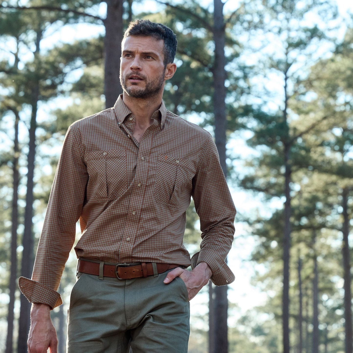 Man in a Tom Beckbe Tidewater Shirt and green pants walking through a forest with tall trees.