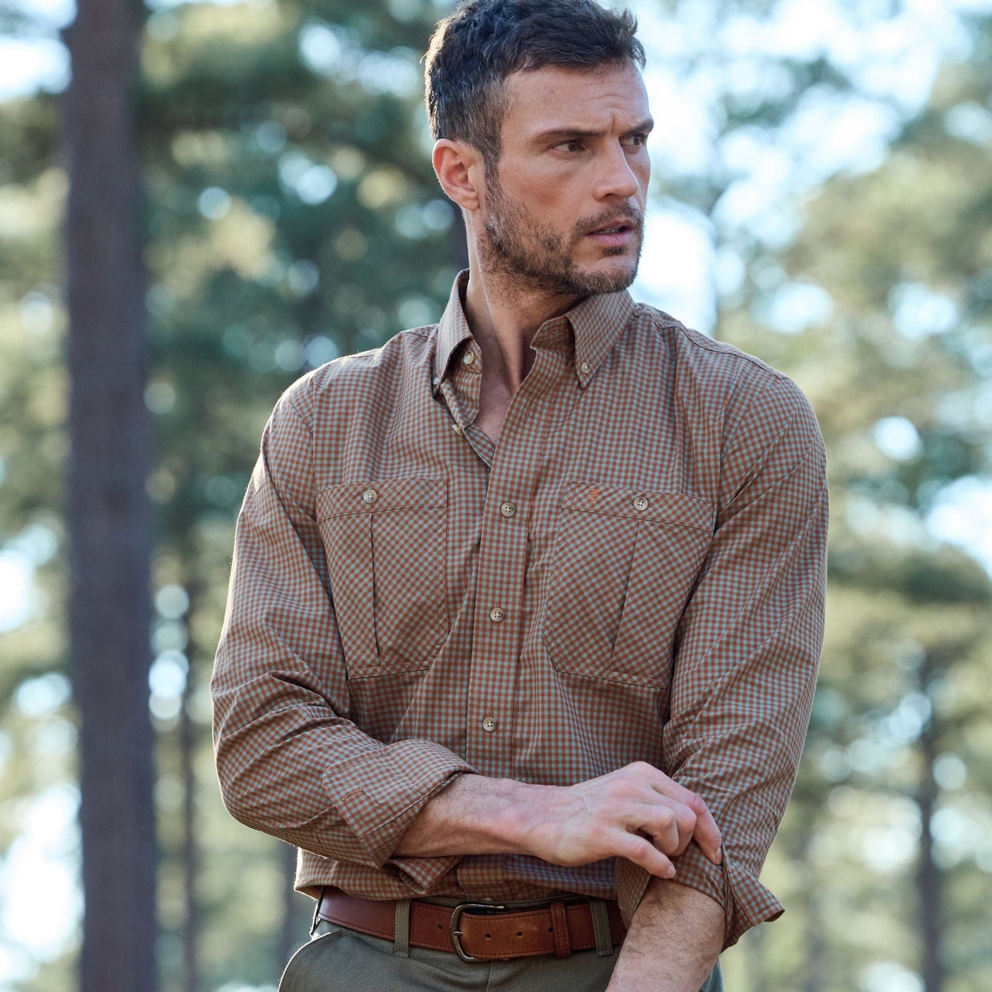Man in a Tom Beckbe Tidewater Shirt rolls up sleeves while standing outdoors among trees with a blurred background.