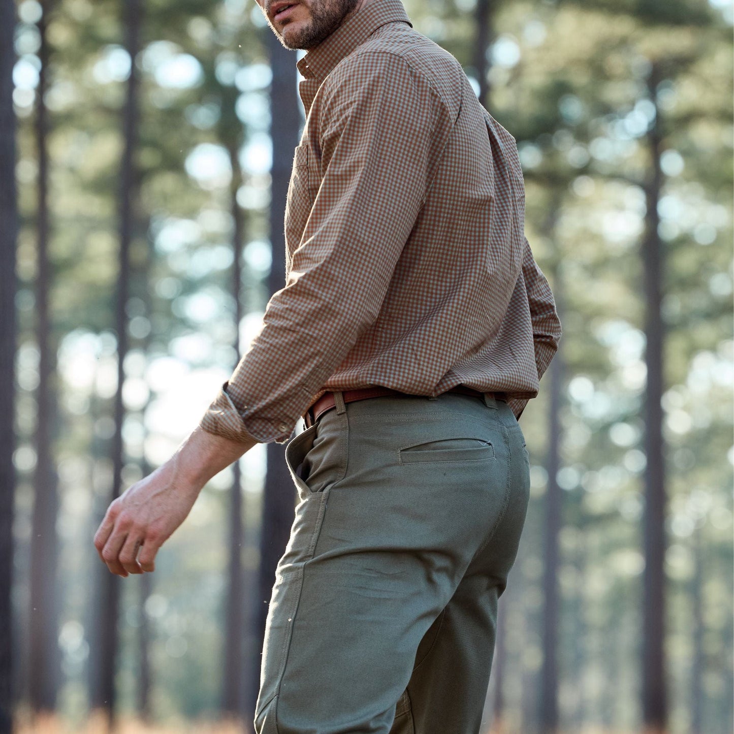 Man in a Tom Beckbe Tidewater Shirt and green pants walking in a forest surrounded by tall trees.