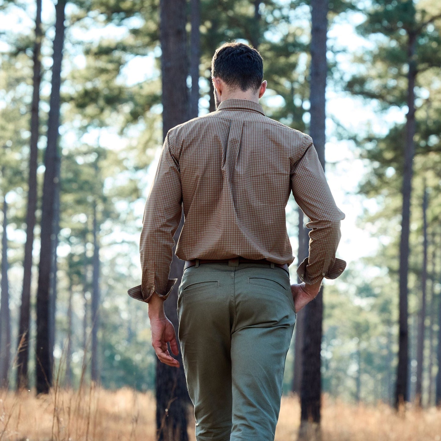 A man in a Tom Beckbe Tidewater Shirt and green pants strolls through a sunlit forest with tall trees.