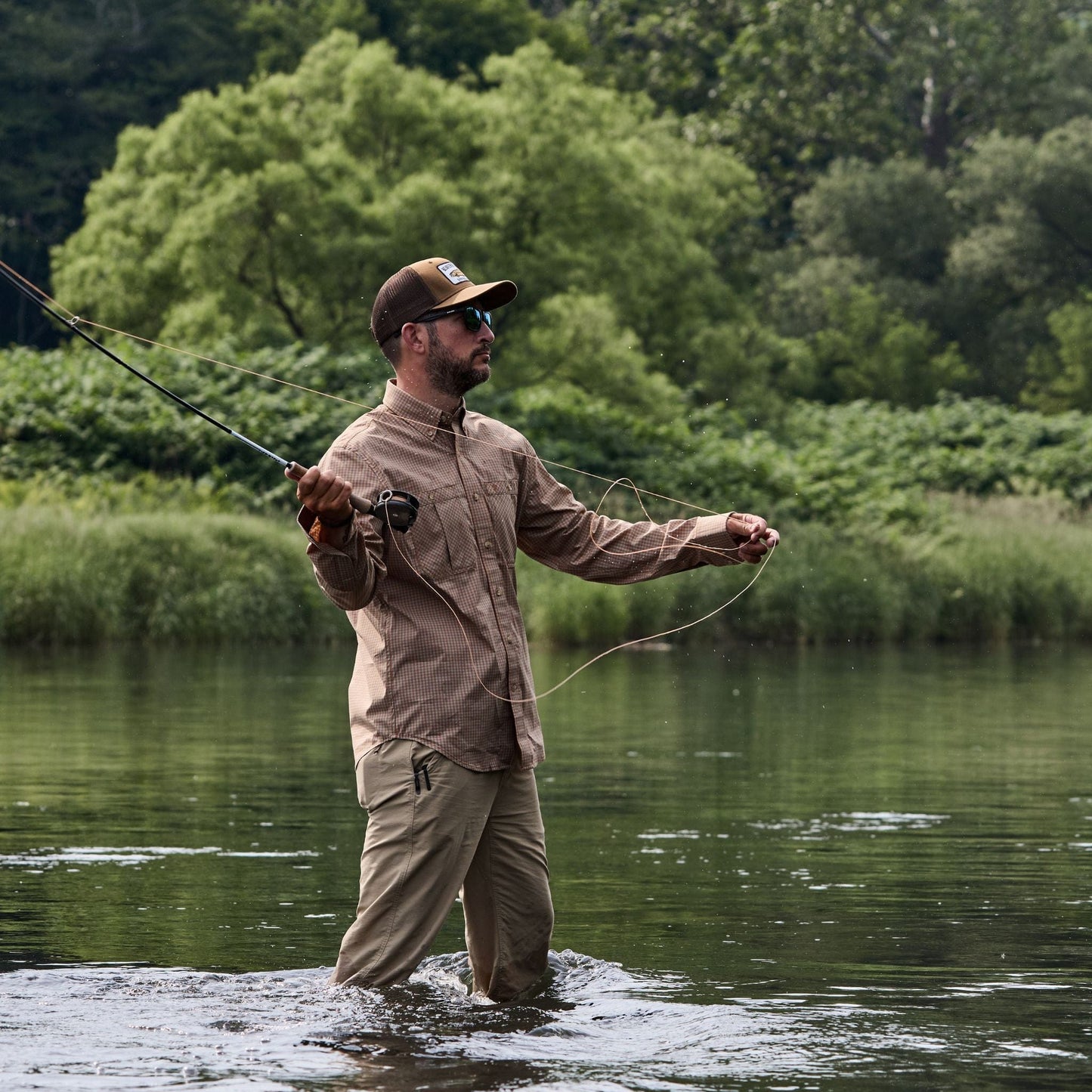 Fly fishing in a river, wearing a Tom Beckbe Tidewater Shirt and khaki pants, with green trees in the background.