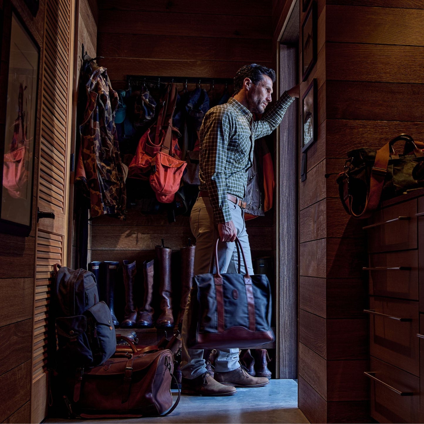 A man in a Tom Beckbe Tidewater Shirt stands in a wooden room with boots, holding a bag, gazing out a small window.