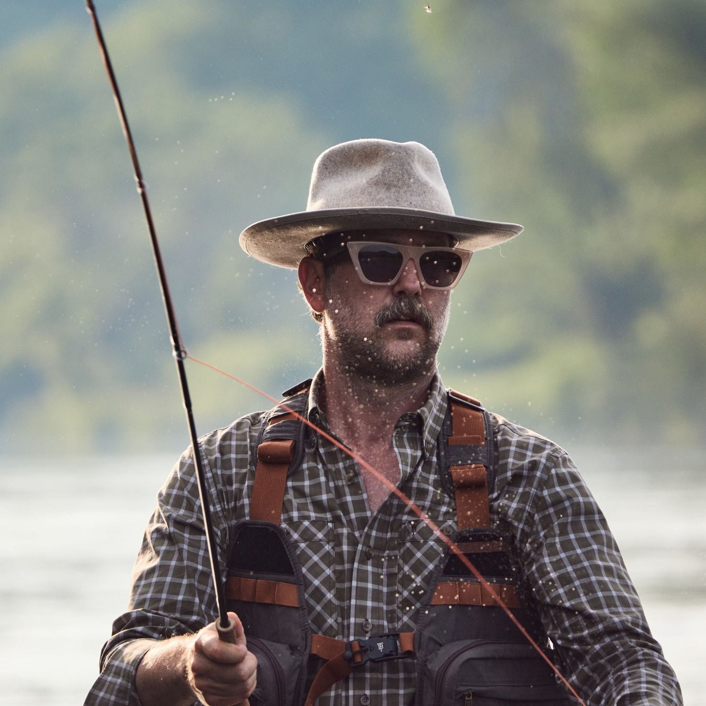 A person in a Tom Beckbe Tidewater Shirt and sunglasses is fishing in a river, rod in hand.