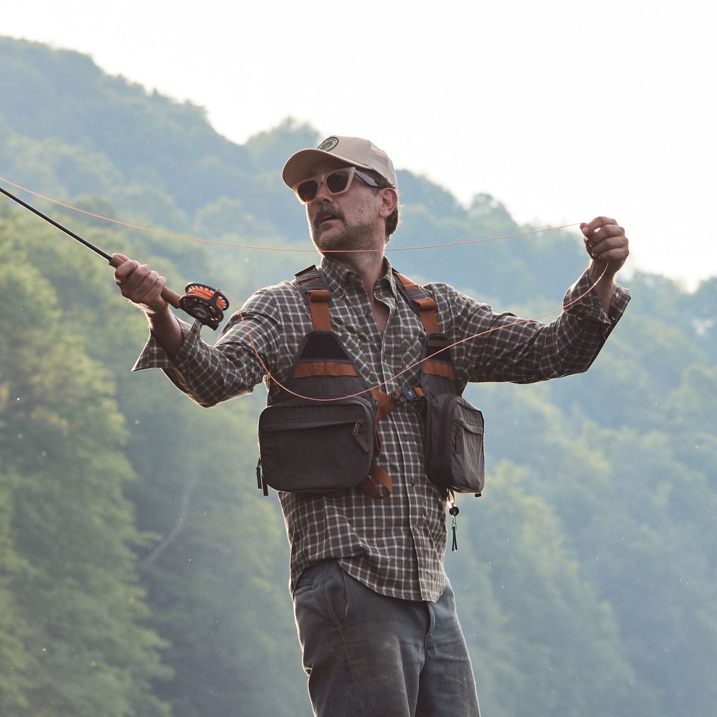 A person in a Tom Beckbe Tidewater Shirt, cap, sunglasses, and holding a fishing rod stands by a tree-covered hill.