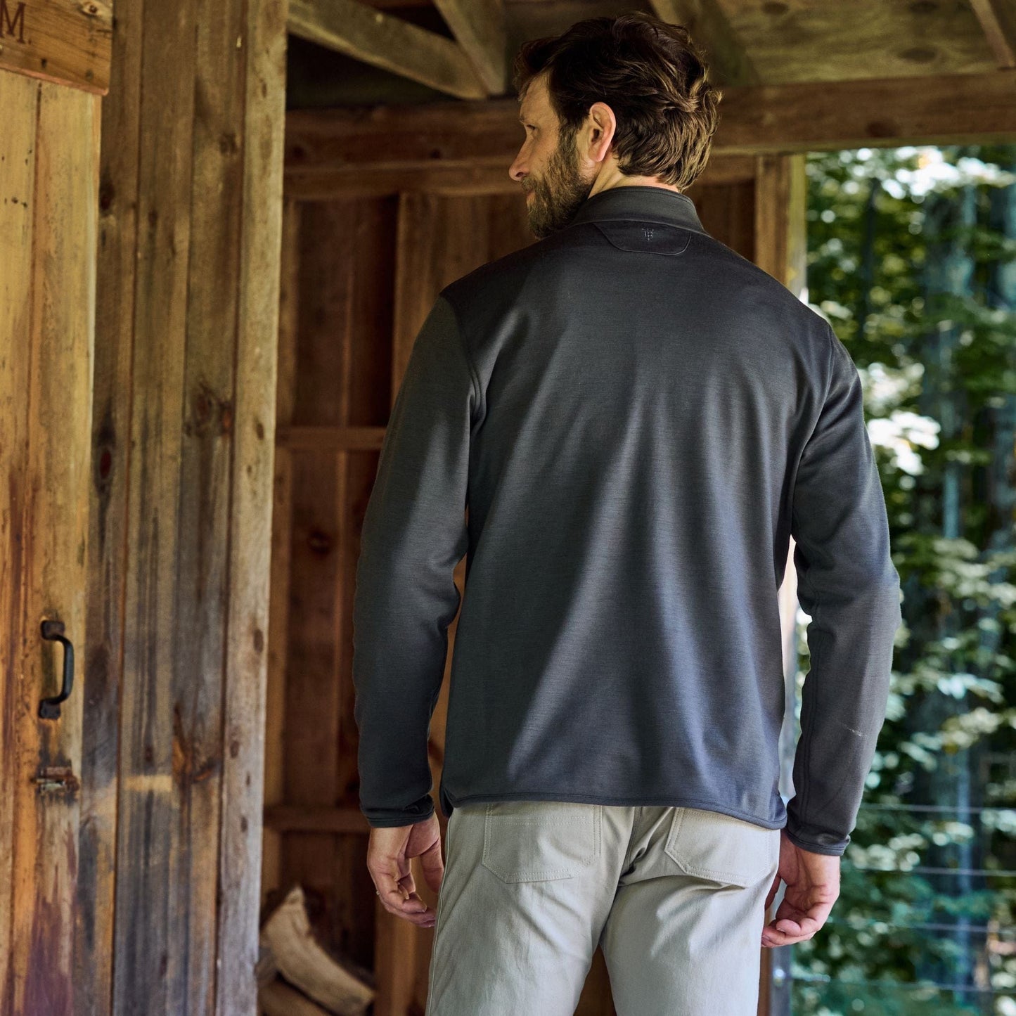 Man in a dark Tucker Fleece Quarter Zip by Tom Beckbe faces away from a rustic cabin, with trees in the background.