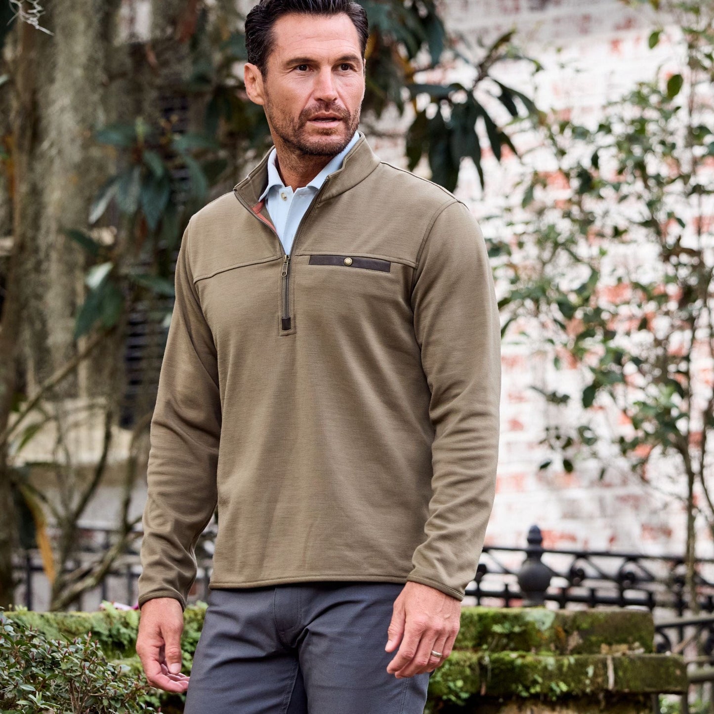 Man in a Tom Beckbe Tucker Fleece Quarter Zip stands outdoors amidst greenery and a brick wall.