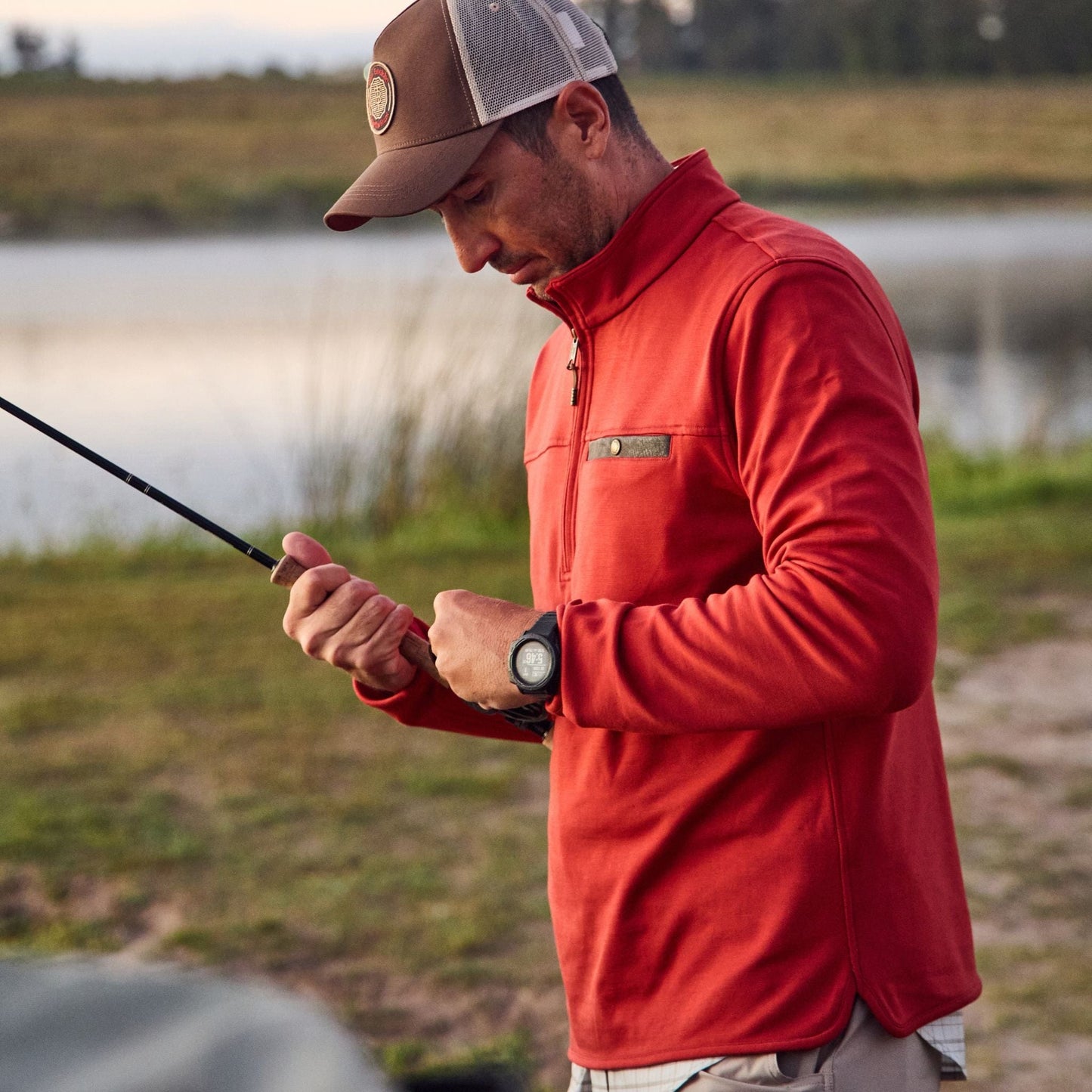 Man in a red jacket and cap, adjusting his line by the lake in his Tom Beckbe Tucker Fleece Quarter Zip.
