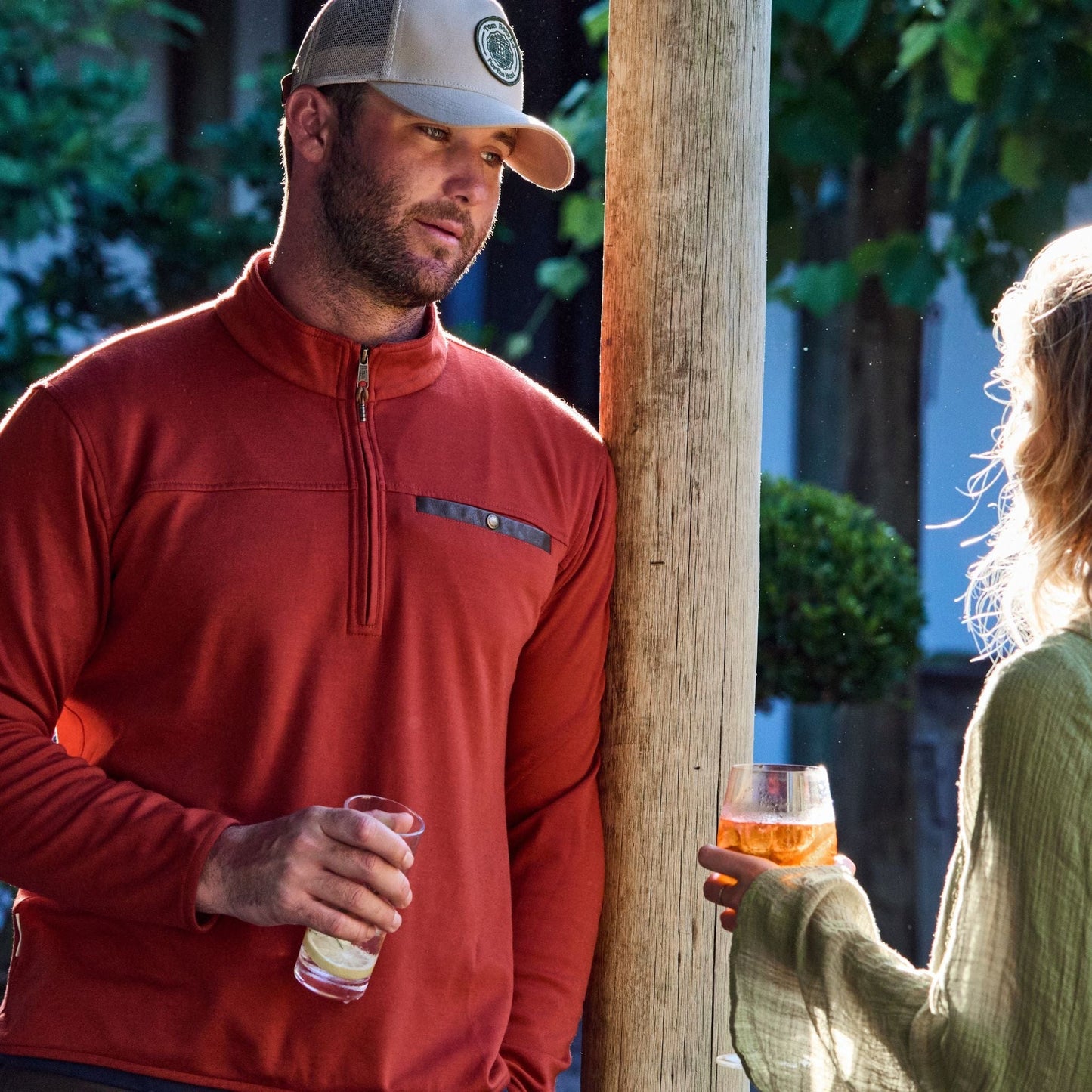 A man in a red Tucker Fleece Quarter Zip by Tom Beckbe chats with a blonde by a wooden post, both enjoying drinks.