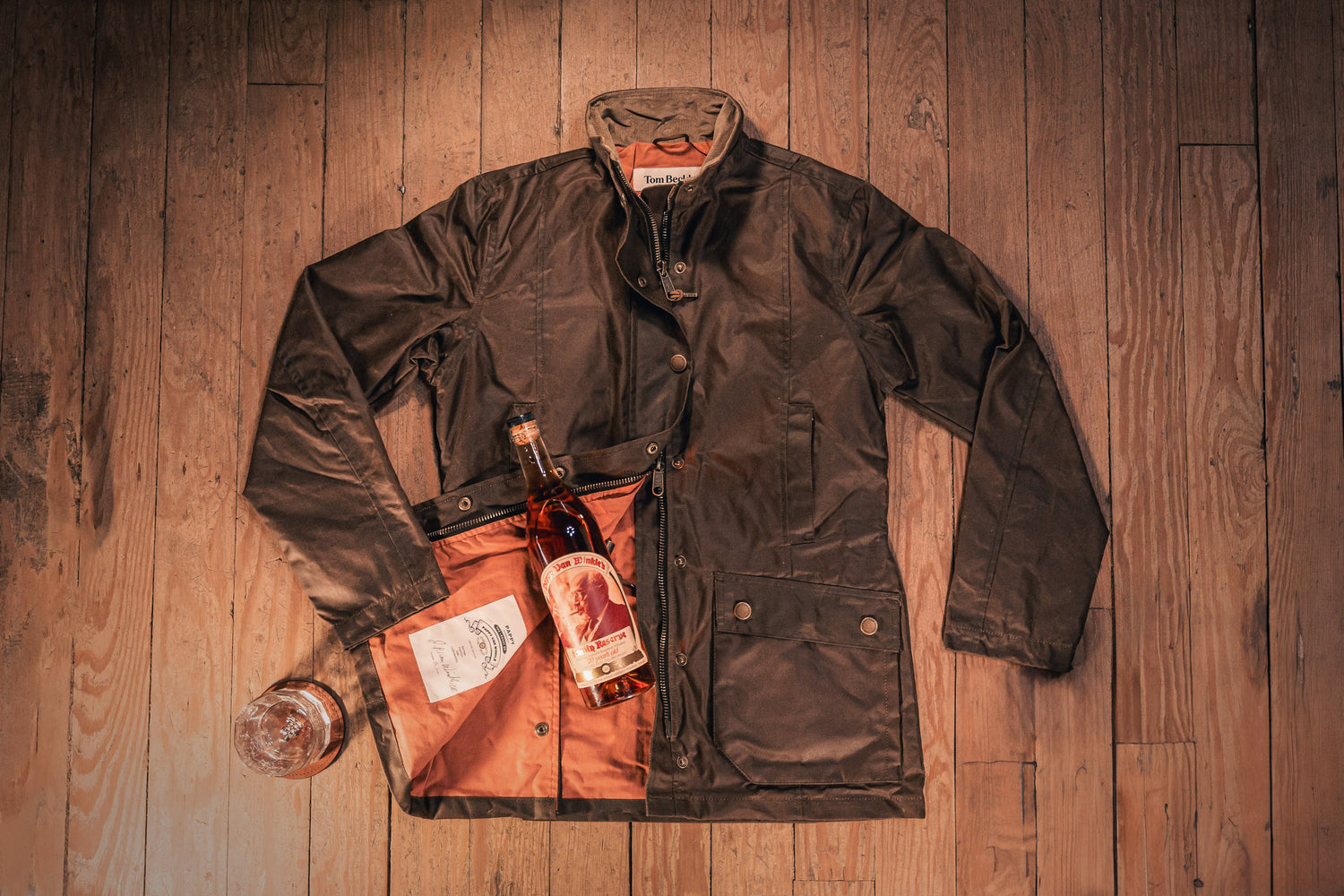 Dark brown jacket on wooden floor with a bottle of whiskey partially placed inside its open pocket. Glass of whiskey nearby.