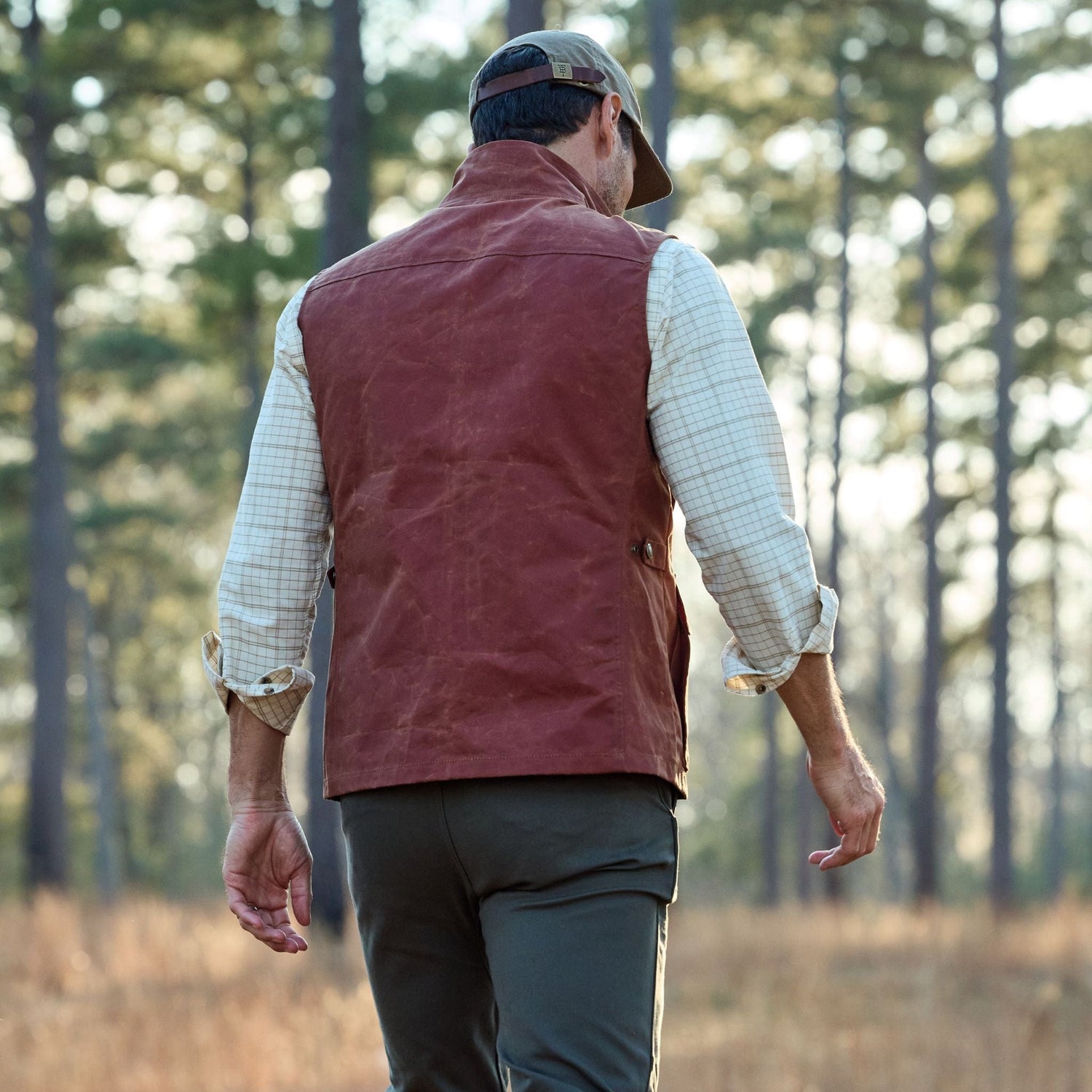 Man in a checkered shirt and brown vest walking through a forest with tall trees.