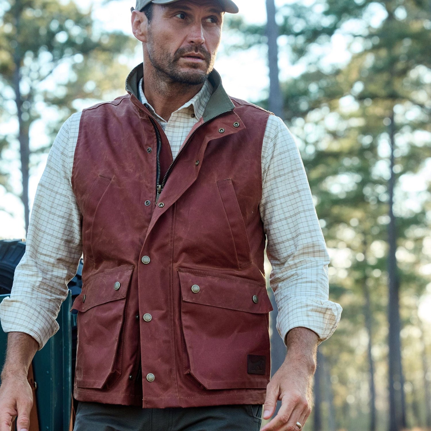 Man in plaid shirt and brown vest stands outdoors with trees in the background.