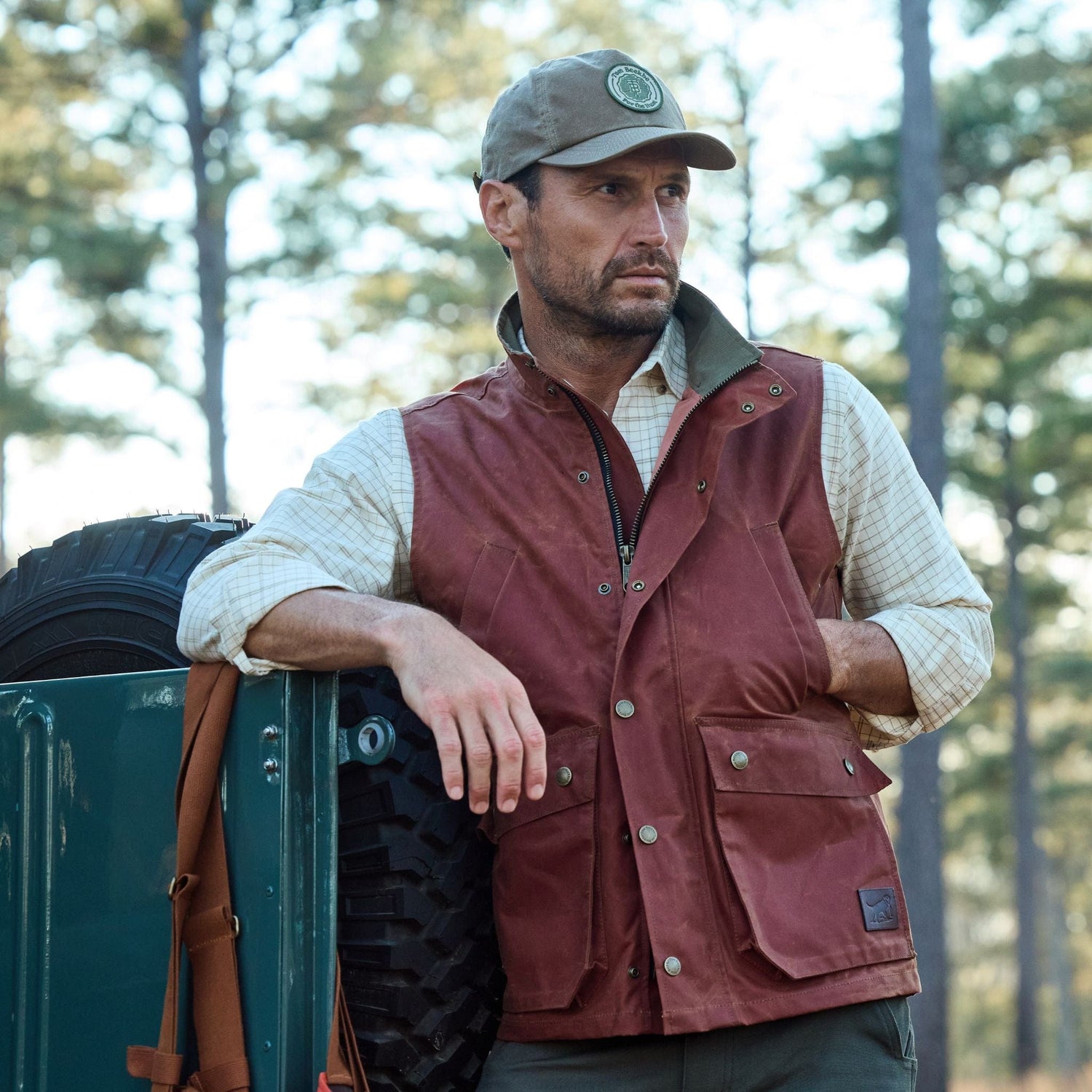 A man in an outdoor setting leans on a vehicle, wearing a cap, red vest, and checkered shirt, with trees in the background.
