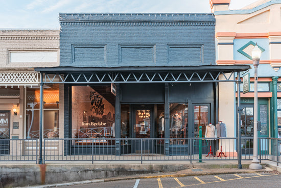 Street view of a dark gray brick storefront with Tom Beckbe signage, glass windows, and clothing displays inside.