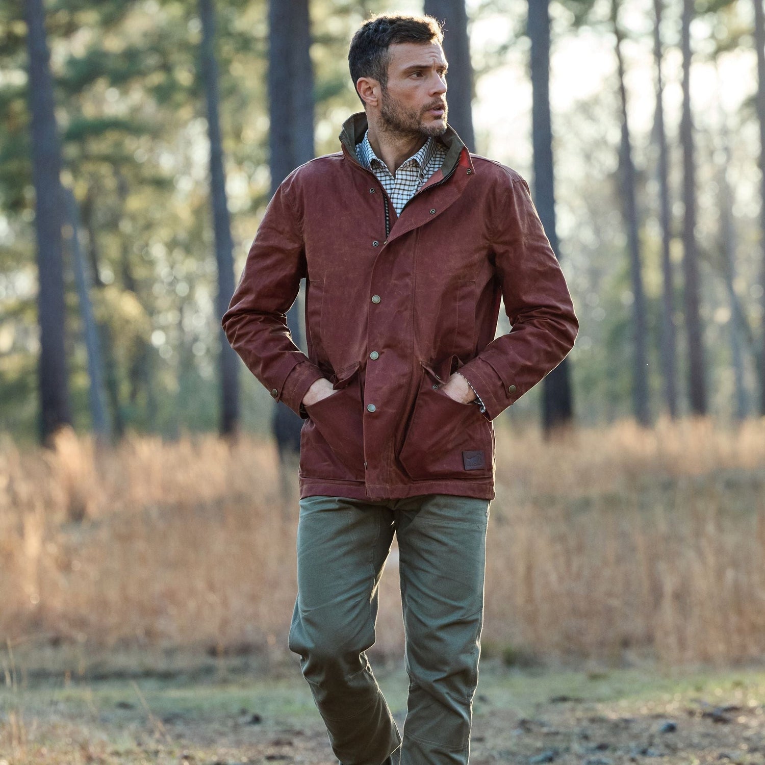 A man in a brown jacket and green pants walks outdoors in a forested area, with trees and sunlight in the background.