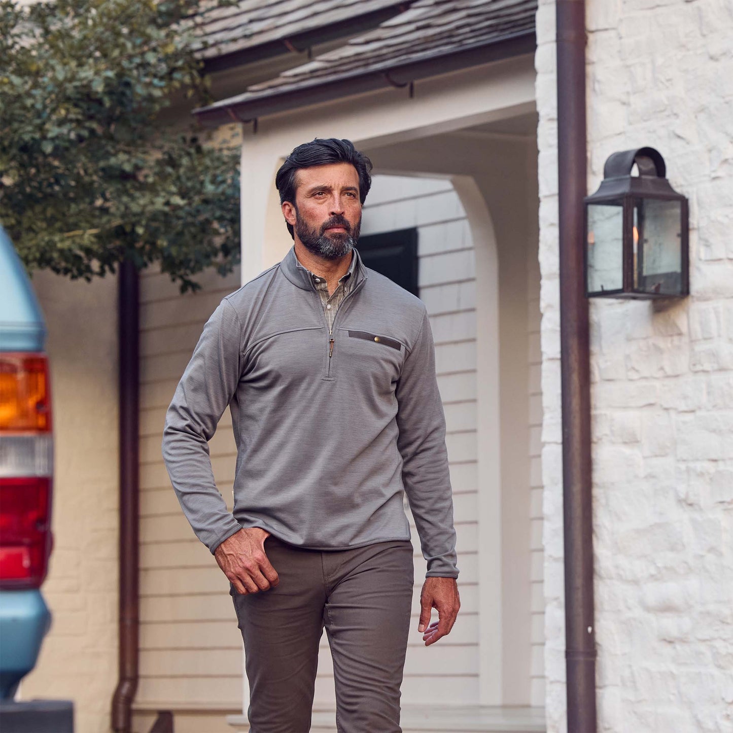 A bearded man walks outside near a white house, wearing Tom Beckbes Tucker Fleece Quarter Zip and brown pants.