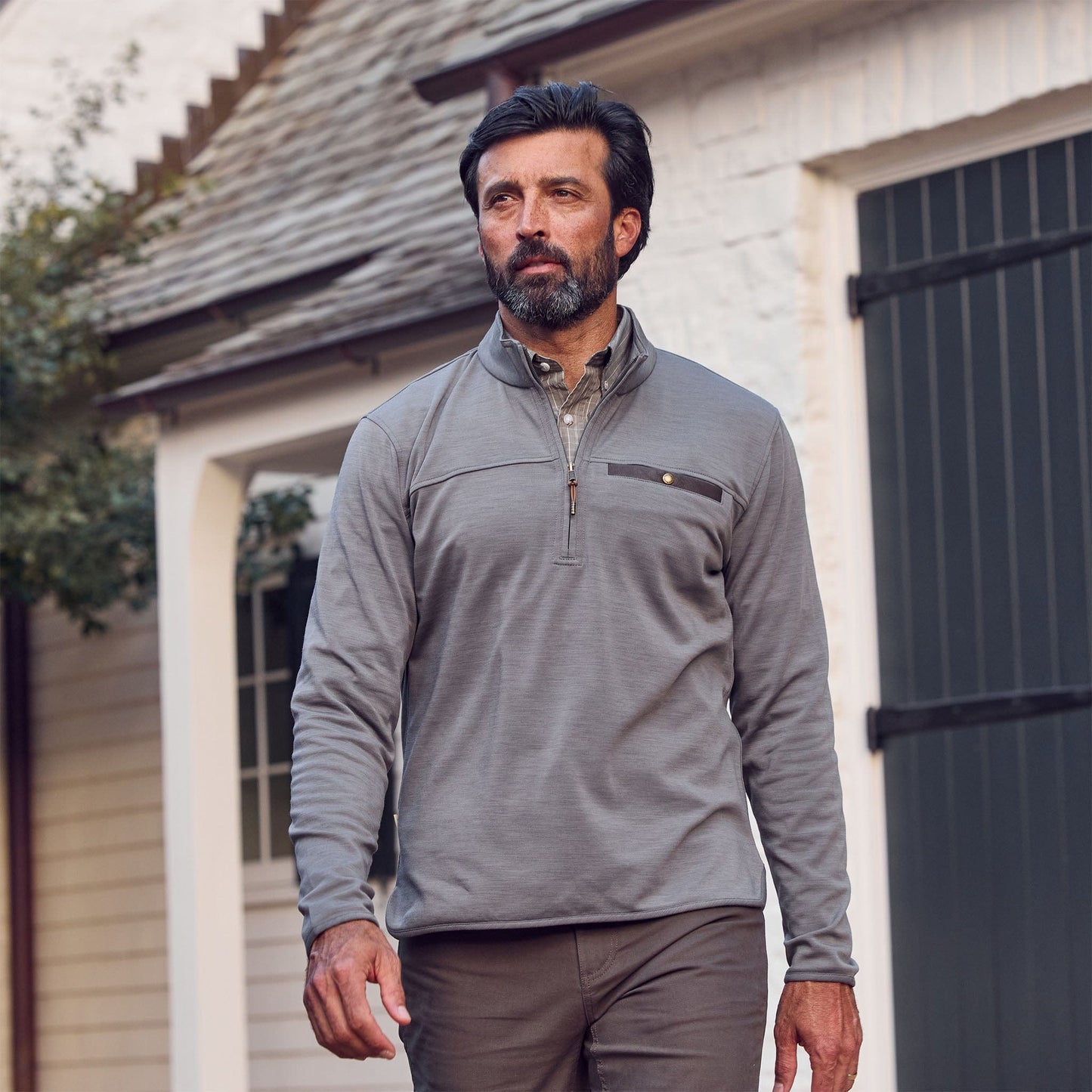 A bearded man with dark hair wears a Tom Beckbe Tucker Fleece Quarter Zip and brown pants outdoors near a house.