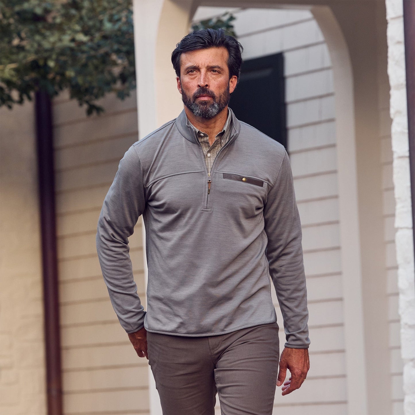 Man with dark hair and beard wears Tom Beckbe Tucker Fleece Quarter Zip and brown pants walking outside by a building.