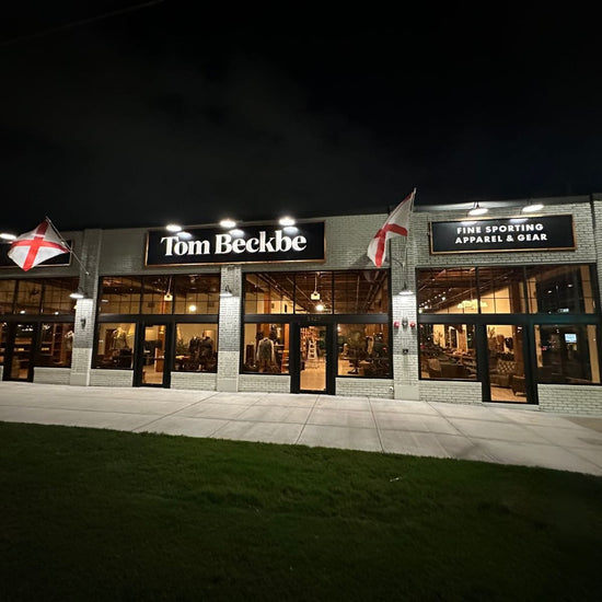 Exterior view of a store named Tom Beckbe, with signs for fine sporting apparel and gear, and flags with red crosses outside.