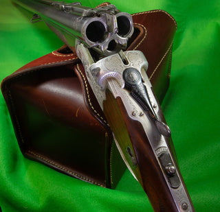 Double-barrel shotgun with ornate engravings, propped on a leather case, on a green background.