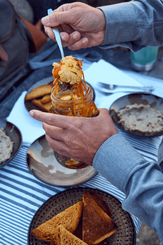 Person scooping vegetables from a jar with a fork at an outdoor table set with triangular toast and plates.