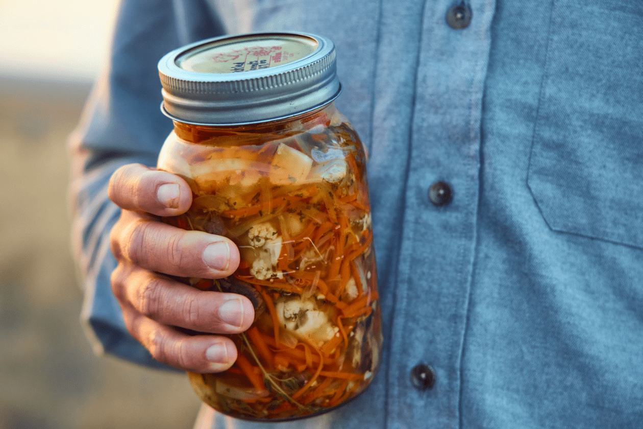 Person holding a mason jar filled with mixed pickled vegetables, including carrots and cauliflower, in front of a chest in a blue shirt.
