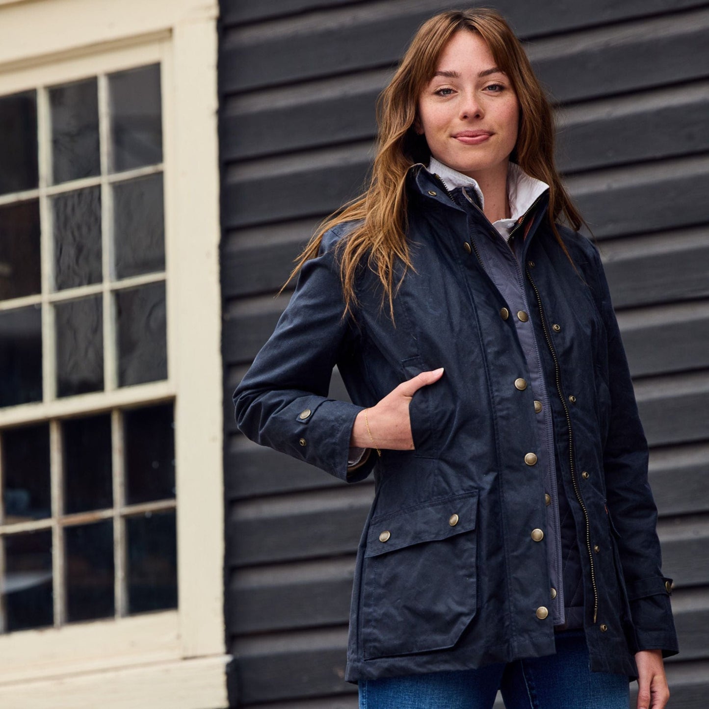 A person wearing a navy jacket stands in front of a dark wooden wall and window, with one hand in a pocket, smiling slightly.