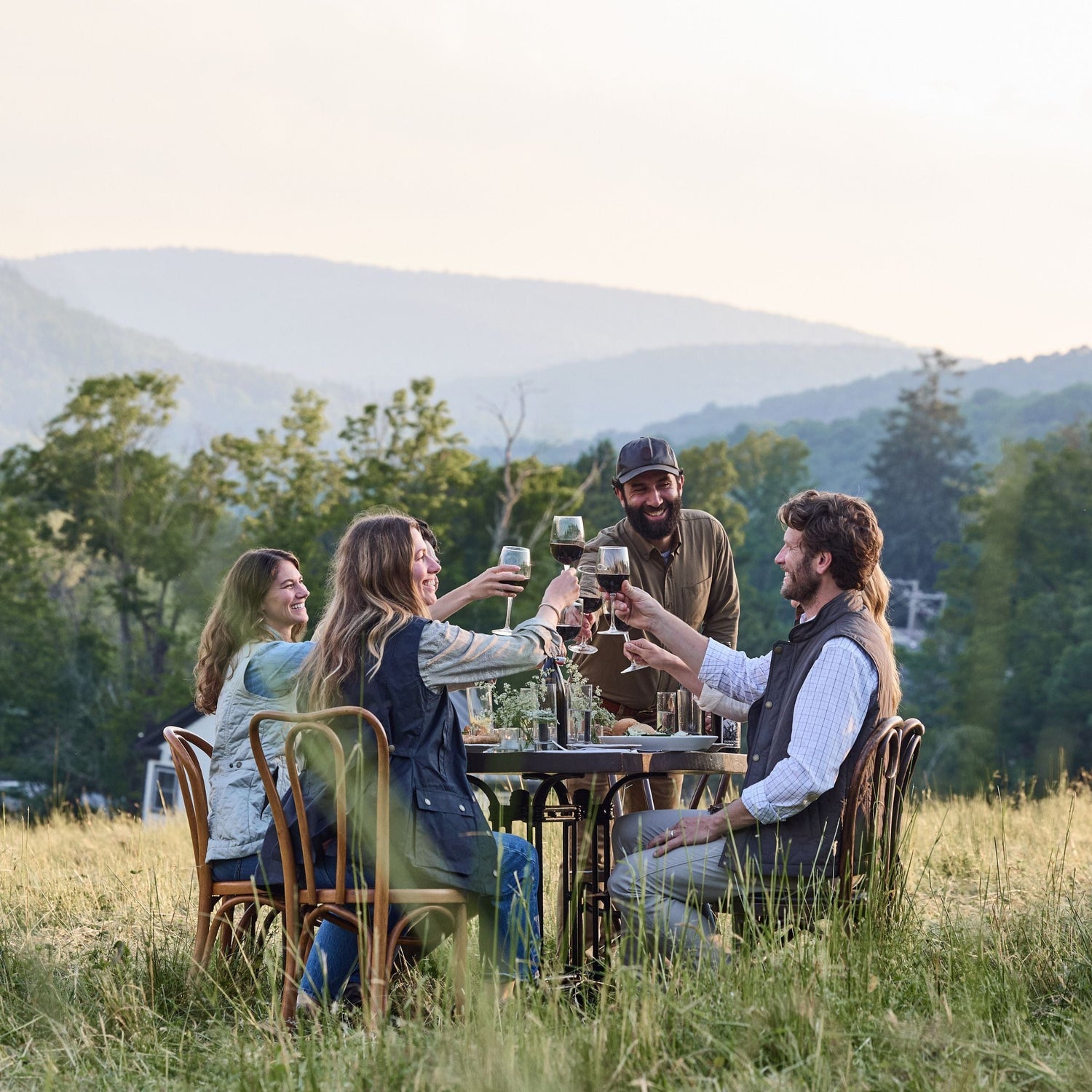 Four people sitting at a table in a grassy field, toasting with drinks, with hills and trees in the background.
