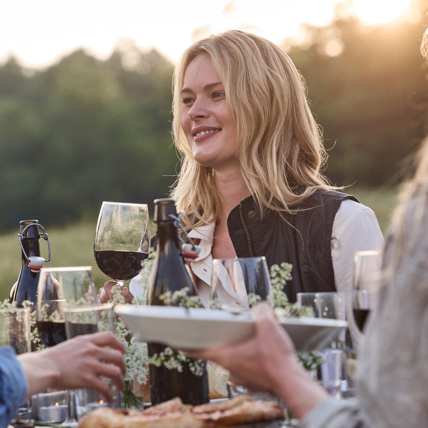 Person sitting at an outdoor table enjoying red wine, surrounded by others, with a blurred background of trees and soft sunlight.