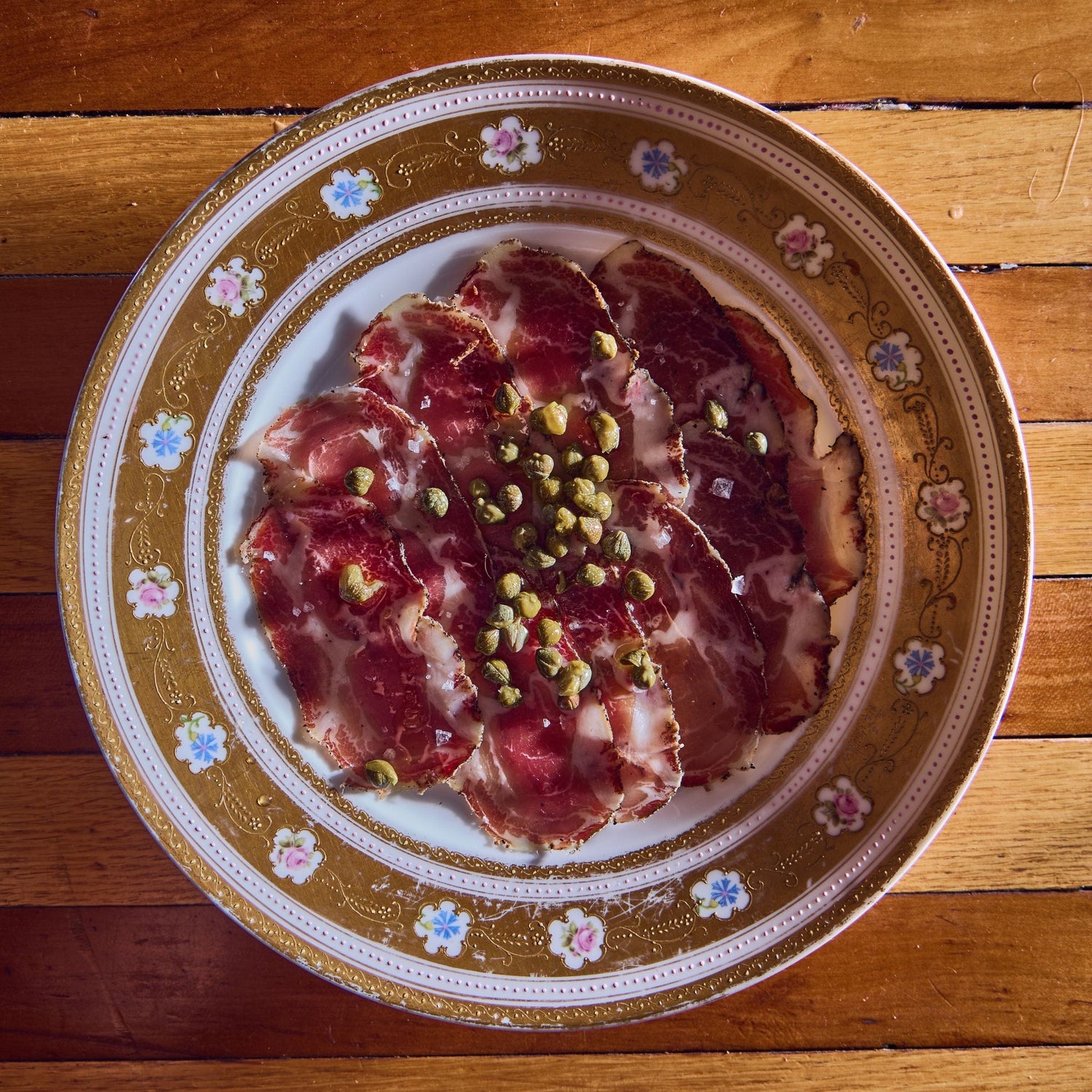 Slices of cured meat topped with capers on a decorative floral plate, set on a wooden surface.