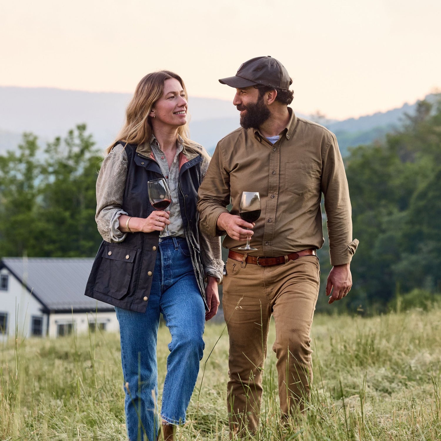Two people walk through a grassy field, holding wine glasses and smiling at each other, with a house and trees in the background.