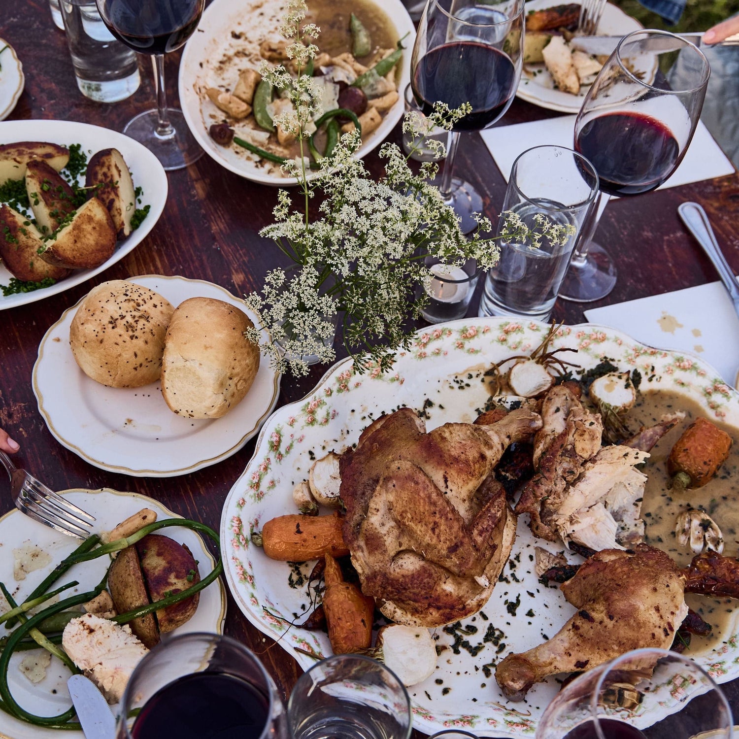 A table set with roast chicken, vegetables, rolls, red wine, and water glasses. White flowers as a centerpiece.