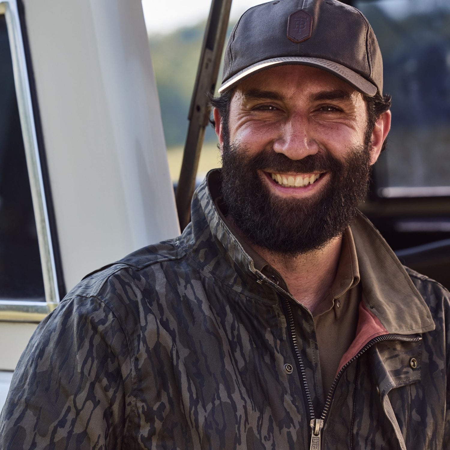 Man with a beard wearing a camouflage jacket and cap smiles outdoors near a vehicle.