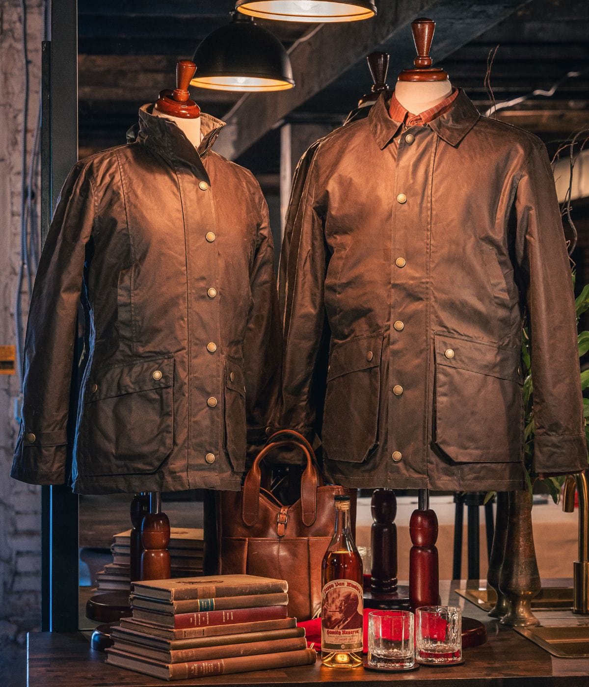 Two brown waxed jackets on mannequins, with vintage books, a leather bag, and bottles on a wooden table under warm lighting.