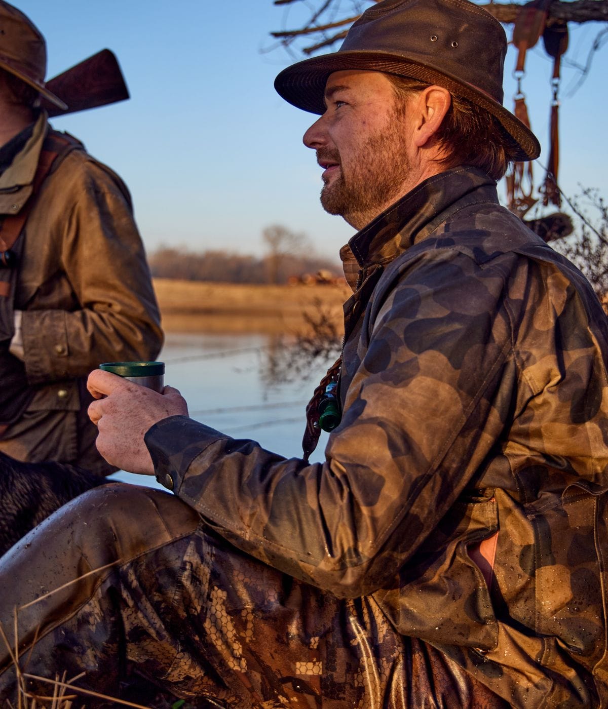 Man in camouflage gear sits outdoors near water, holding a mug. Another person in similar attire is partially visible.