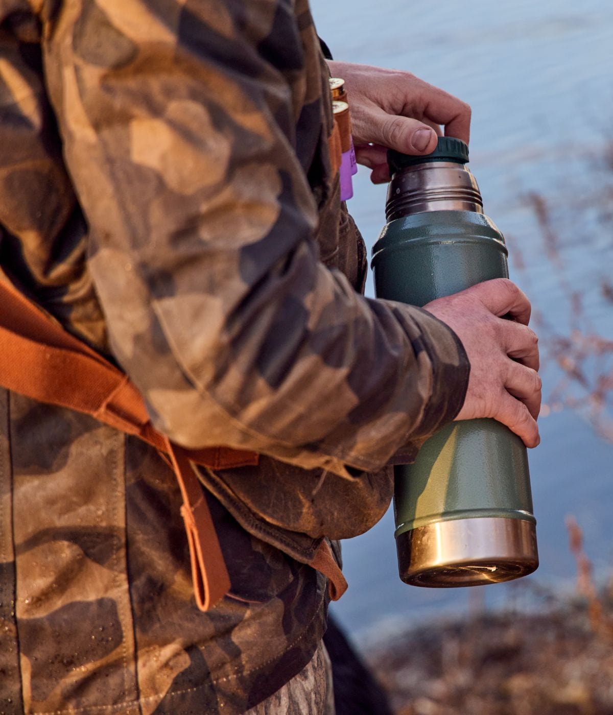 Person in camouflage jacket holding a large green thermos near a body of water.