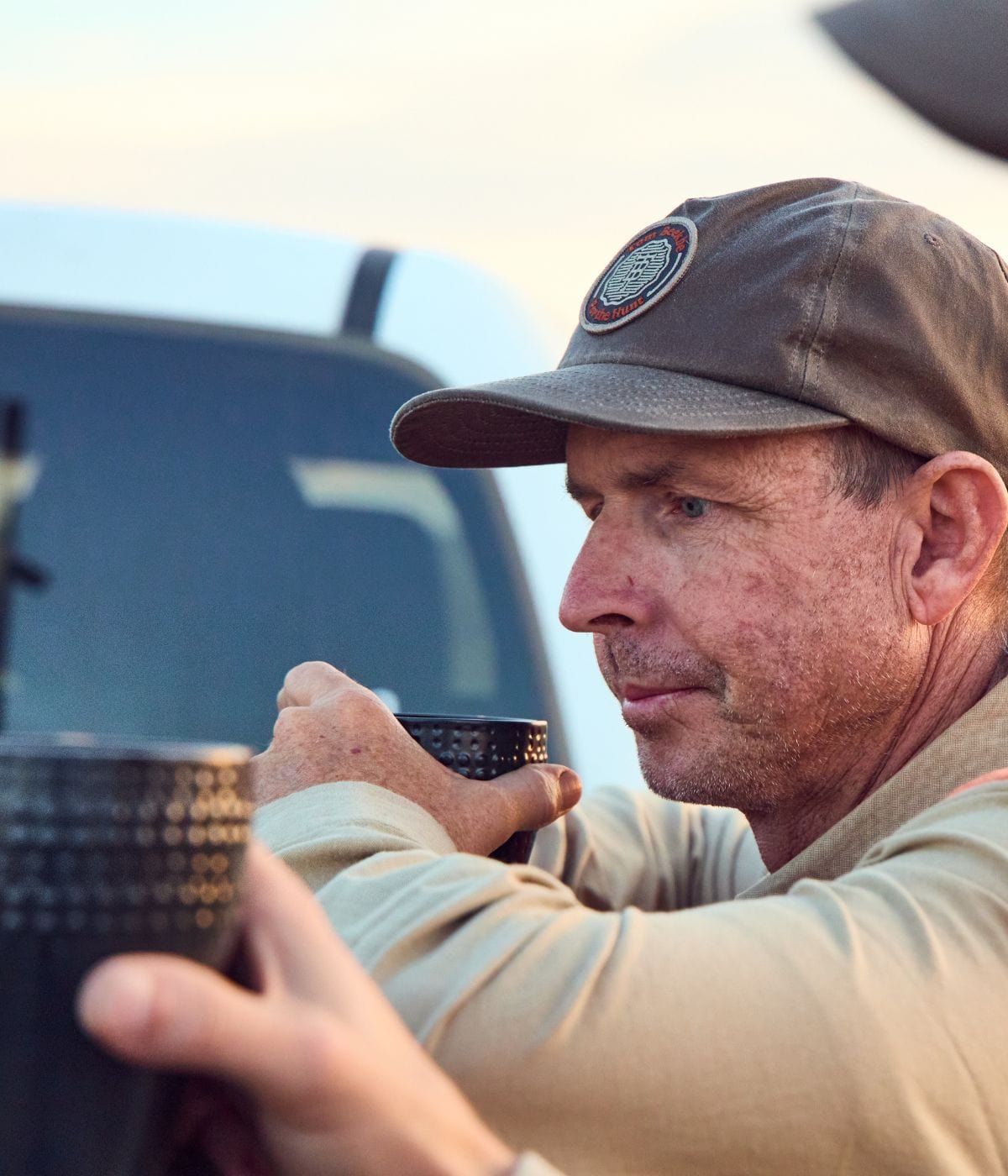 Man in a cap holding a textured cup, leaning against a vehicle, with a neutral expression.