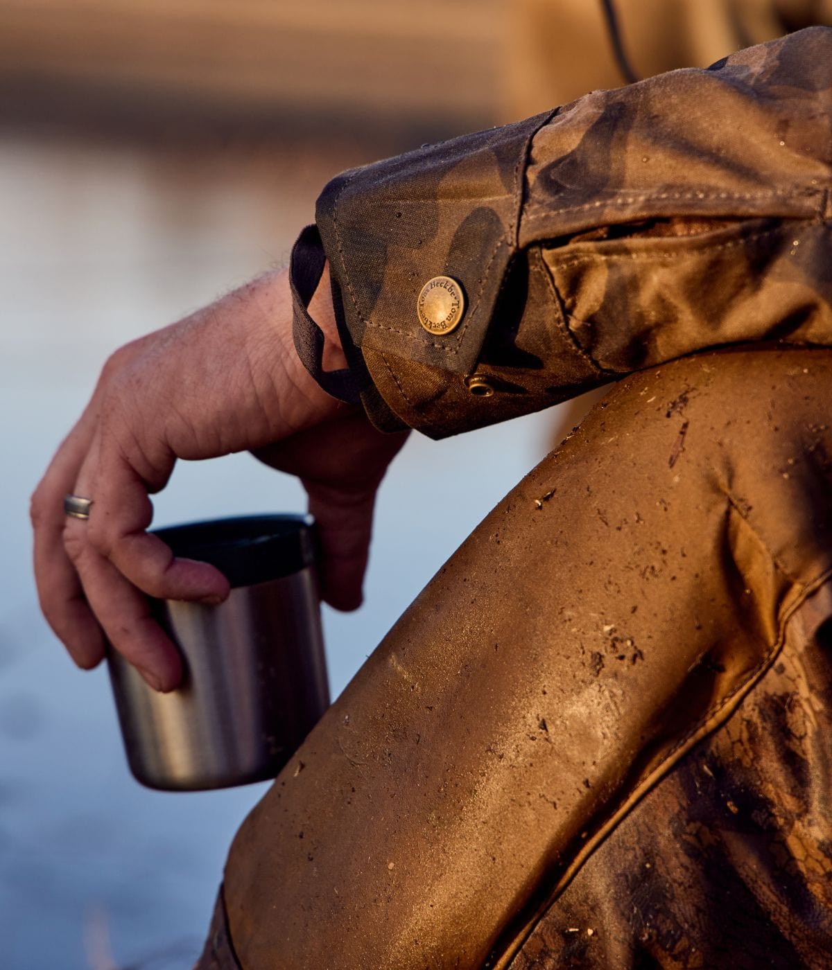 Person wearing muddy waterproof clothing holds a stainless steel cup by a body of water, with a visible ring on their finger.