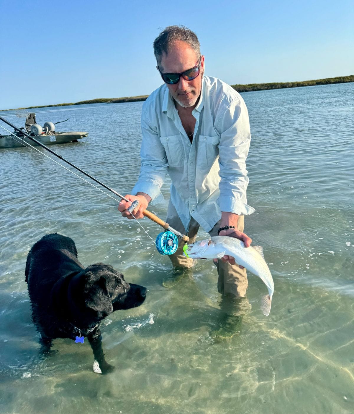 Man holding a fish with a fishing rod in shallow water, standing next to a black dog, under clear blue sky.