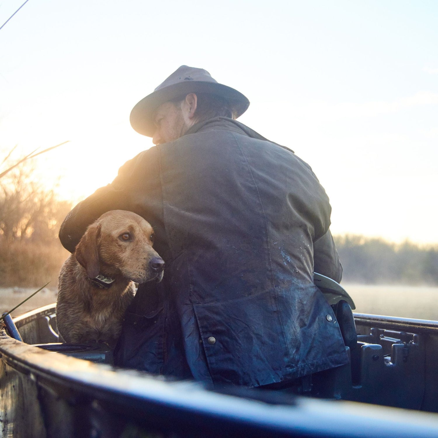 Person with a hat sitting in a boat on a calm waterway at sunrise, accompanied by a dog.