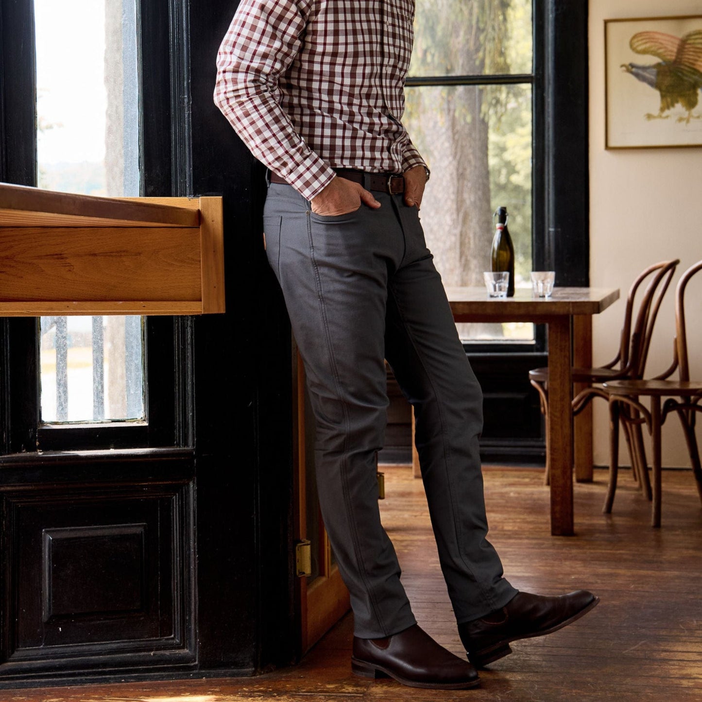 A person in a plaid shirt and Tom Beckbes Redmont Pant leans on a wooden windowsill near a wine bottle.