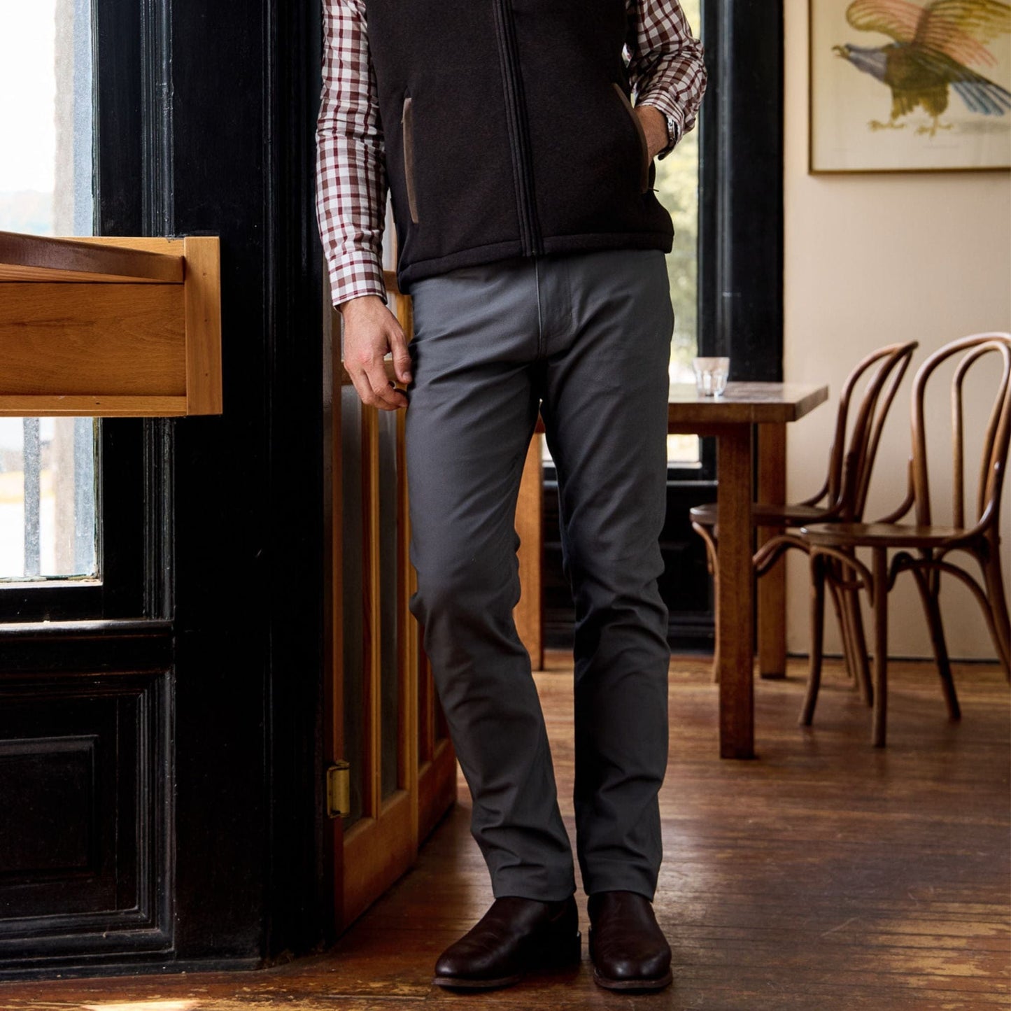 Person wearing a plaid shirt, dark vest, Redmont Pant by Tom Beckbe, and brown shoes; dining area partly visible.