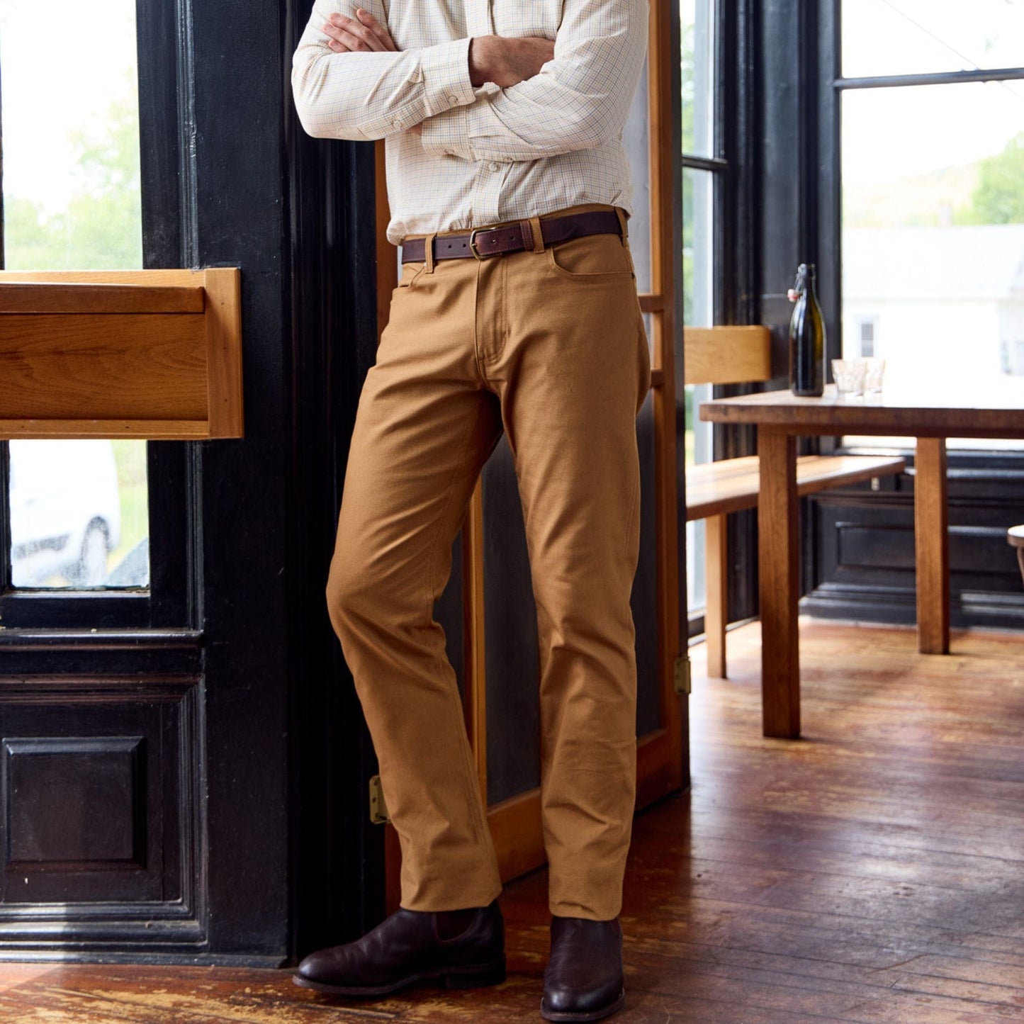 Someone in a light shirt and Tom Beckbes Redmont Pants stands with arms crossed in a cafe with large windows and wood floors.