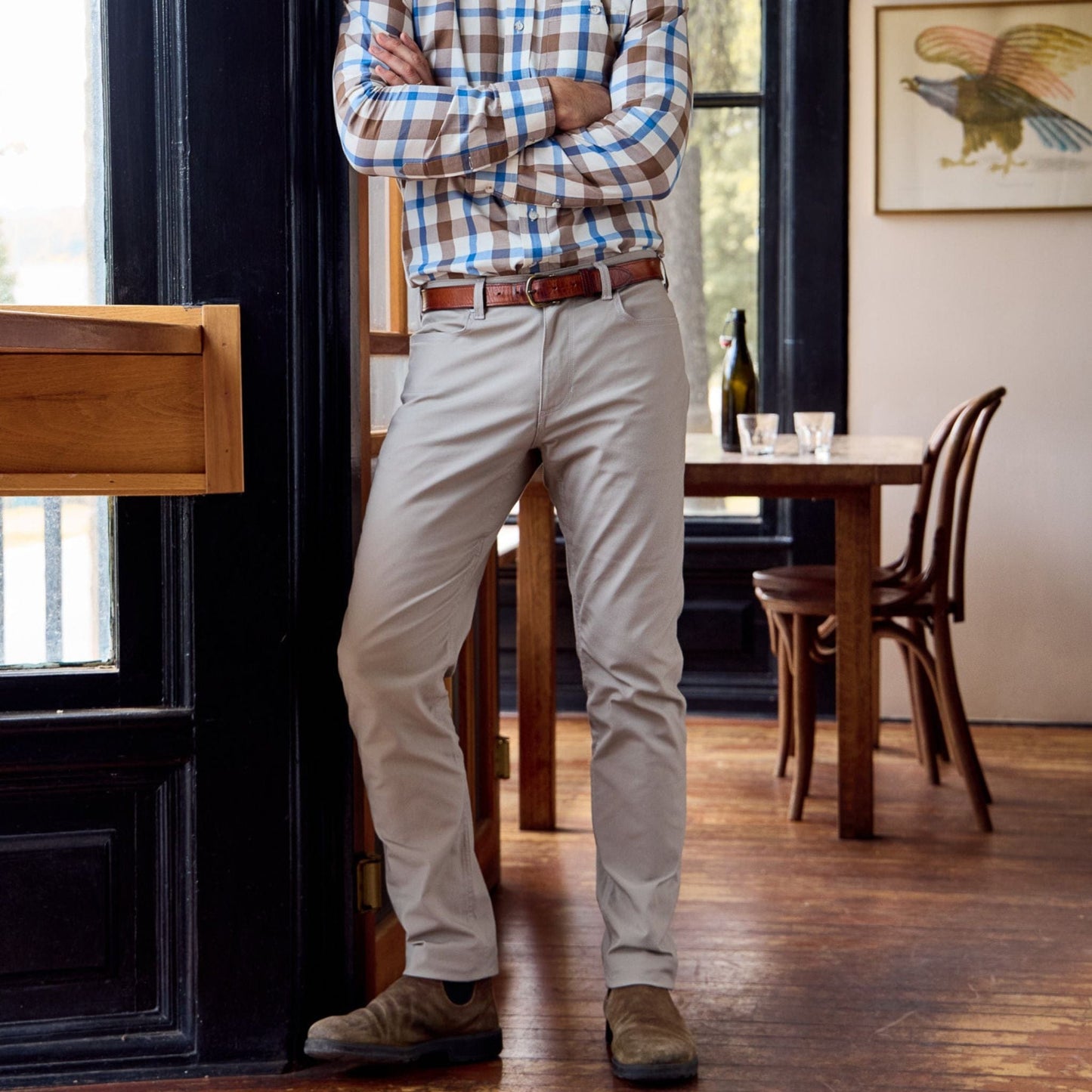 A person in a plaid shirt and Tom Beckbes Redmont Pant stands in a room with wooden floors, table, chairs, and framed art.